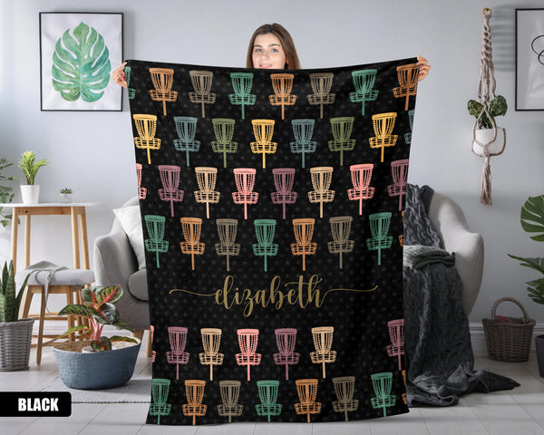 Person holding a black blanket with colorful disc golf basket pattern and name text in a room.