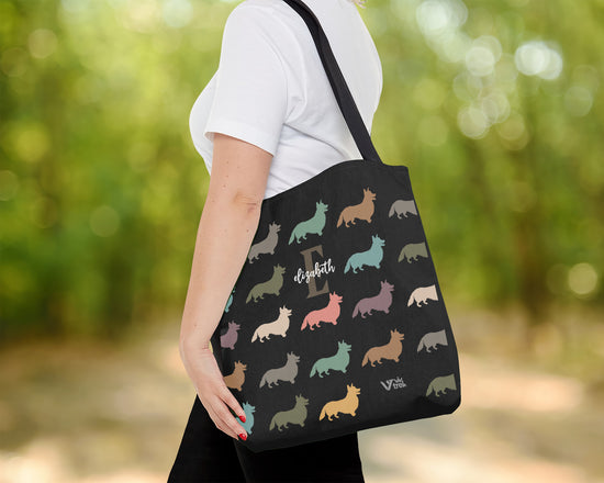 Person holding a black tote bag with colorful dog pattern and initial name outdoors