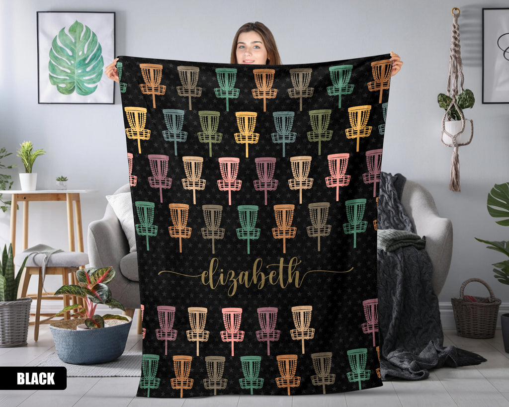 Person holding a black blanket with colorful disc golf basket pattern and name text in a room.