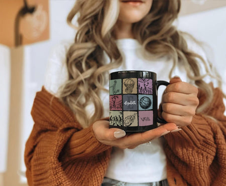 Person holding a black mug with colorful designs and initial name text.