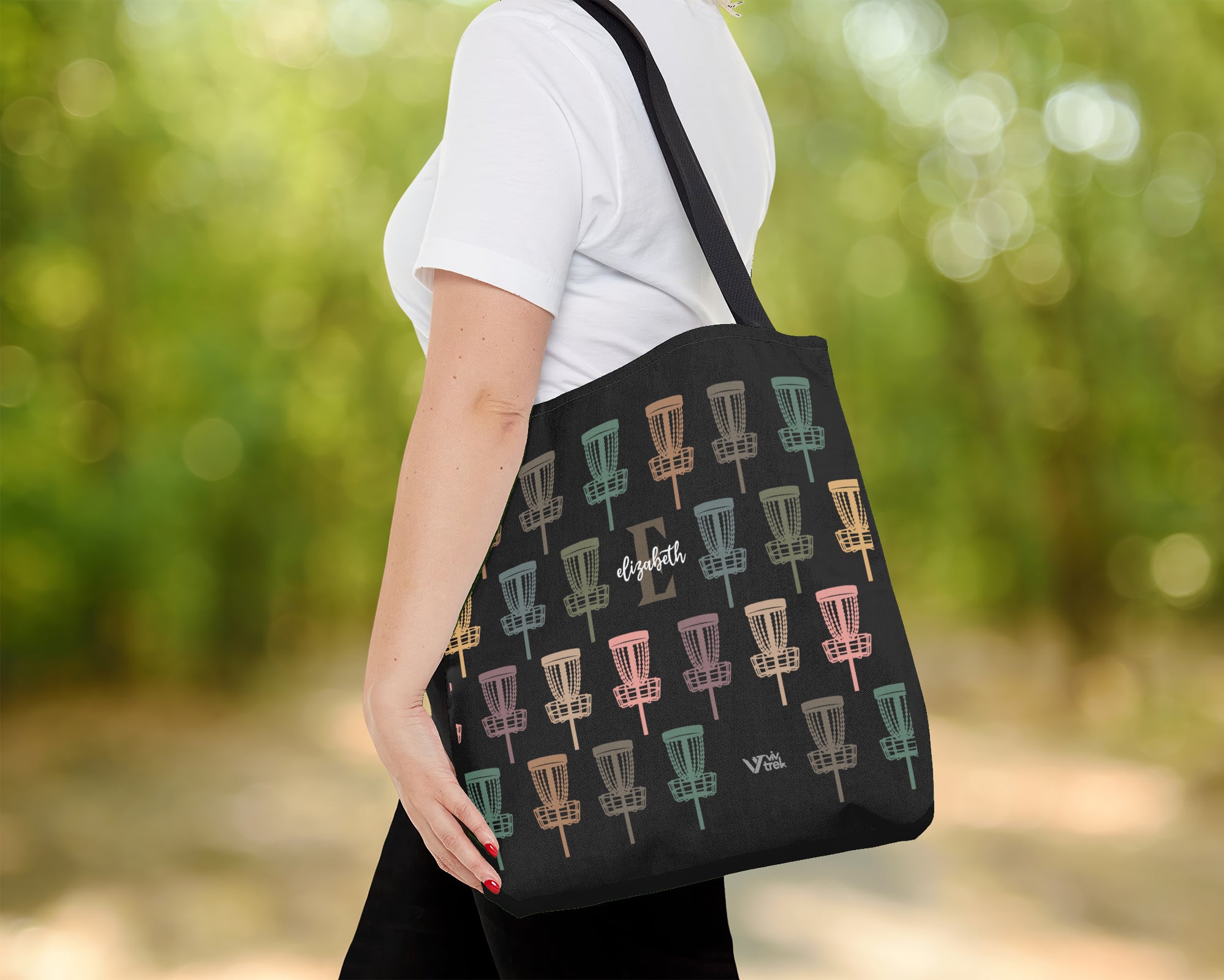 Person holding a black tote bag with name text colorful disc golf disc pattern outdoors.