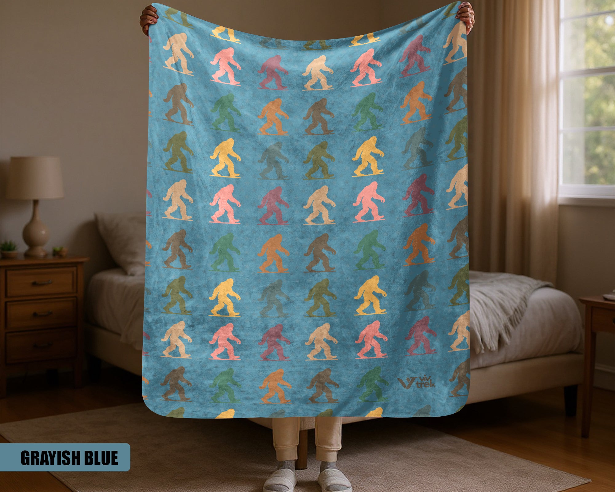 Person holding a colorful 'Bigfoot' patterned blanket in a room with a bed and lamp.