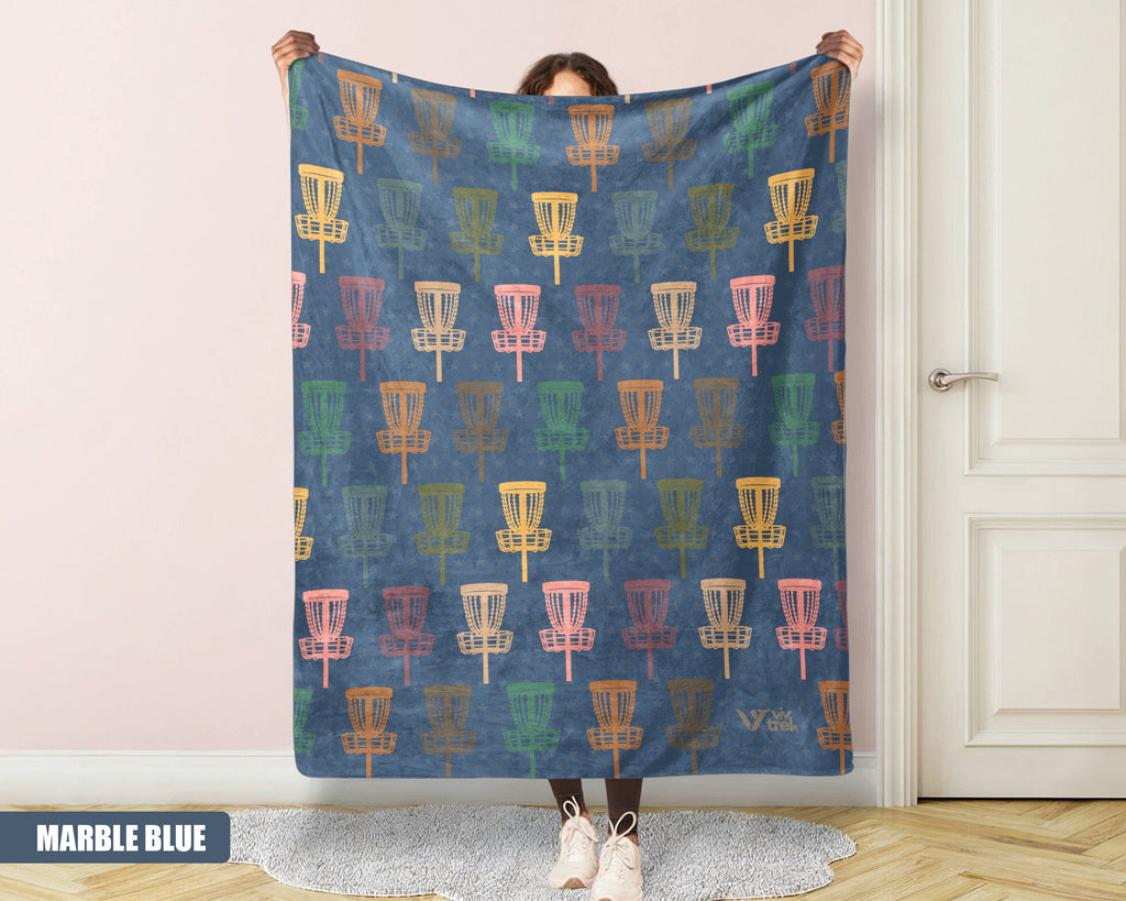 Person holding a colorful patterned blanket with disc golf basket design against a white wall.