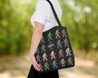 Person holding a tote bag with colorful bigfoot silhouettes and initial name printed on it, against a blurred green outdoor background.